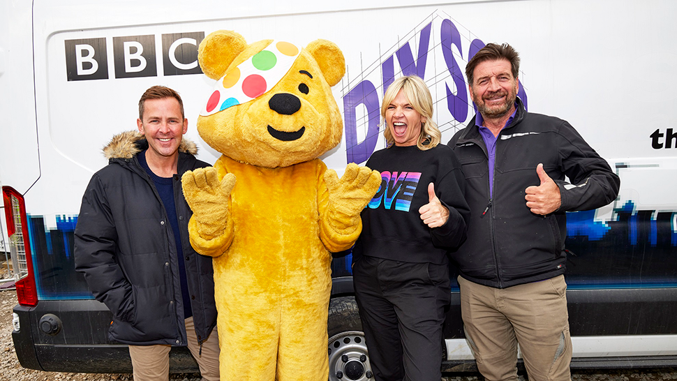 Team of smiling celebrities with BBC Children in Need mascot in front of DIY SOS van