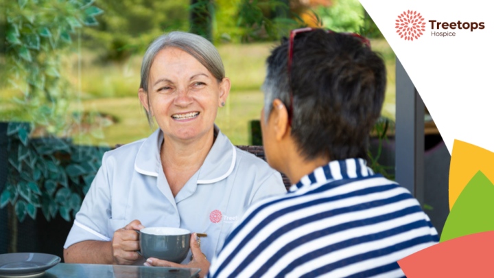 Two women sitting at a table on a shady patio, chatting and drinking tea. One is a Treetops Hospice Healthcare Assistant, wearing a grey nurse uniform. The other is a patient.