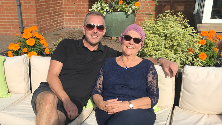 Mum and son sitting on a white outdoor sofa both wearing sunglasses and smiling at the camera