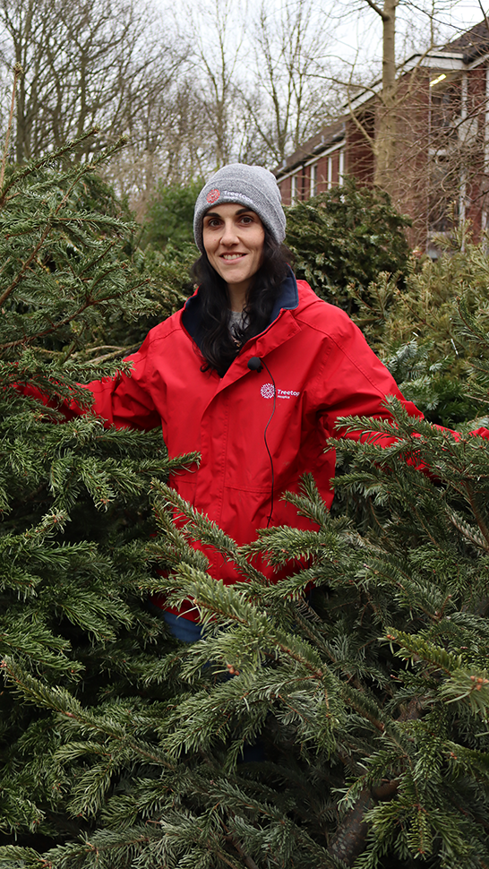 White woman with long black hair dressed in Treetops red coat standing amongst real Christmas trees