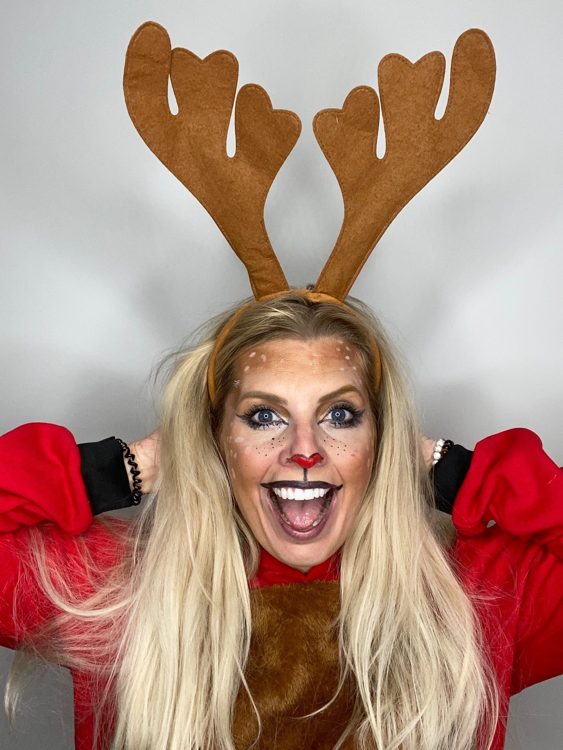 Young white woman with long blond hair wearing reindeer antlers and face make up, smiling with hands behind her head
