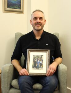 Man with a beard sitting in armchair holding a photo of his mum