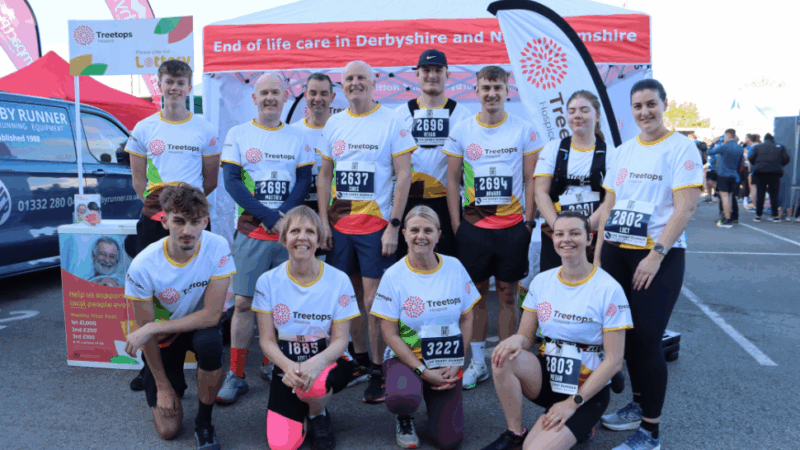 A group of runners in Treetops branded shirts pose together before a charity event, with banners promoting end-of-life care in the background.