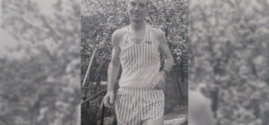 A black and white photo of a man in striped athletic wear standing outdoors.