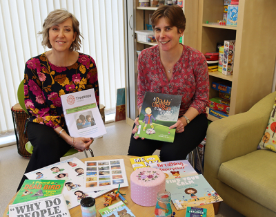 Two women sitting with books in a room full of children's toys and furniture.