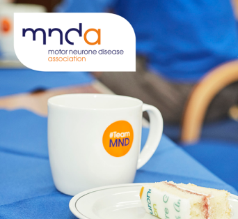 A mug with the Motor Neurone Disease Association logo beside a slice of cake on a blue tablecloth.