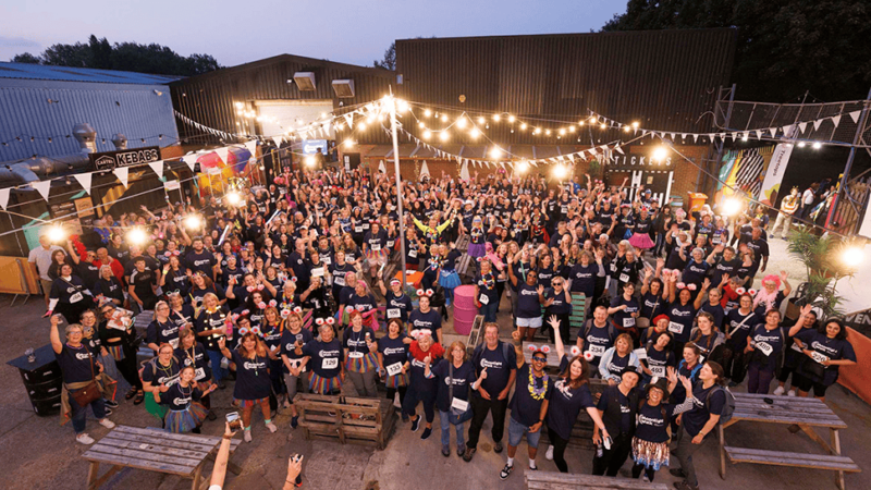 A large outdoor gathering of people smiling and waving at the Treetops Moonlight Walk event with string lights at dusk. Photo credit: Richard Richards Photography