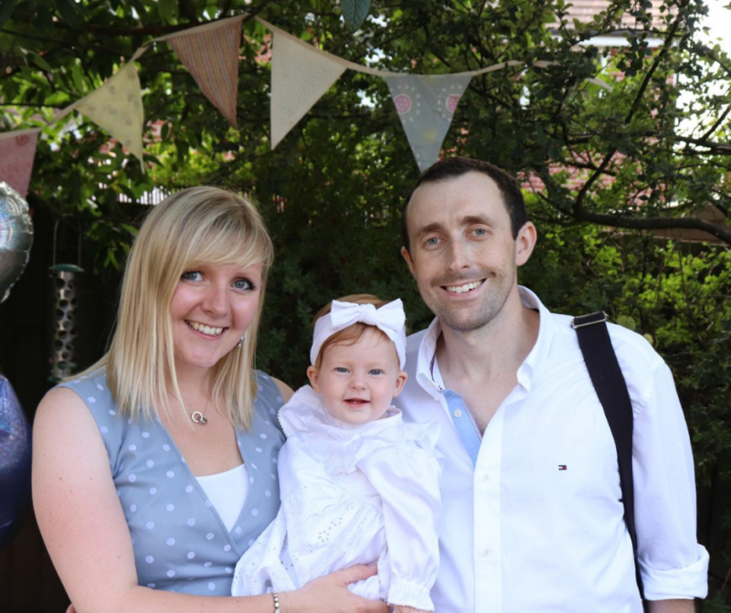 A family stands together outdoors, smiling. The mother in a polka dot dress holds a baby dressed in white, while the father stands beside them.