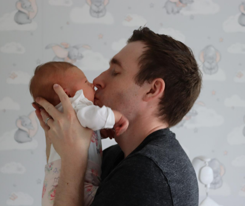 A parent gently holds a baby feeding from a bottle, surrounded by a whimsical nursery wall featuring elephants and clouds.