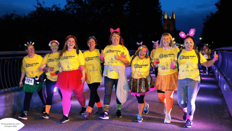 A group of excited participants in yellow shirts, colourful costumes, and accessories walk together at night during a charity event.