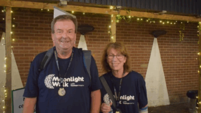 Two individuals wearing matching "Moonlight Walk" t-shirts stand together in front of a brick wall adorned with fairy lights.