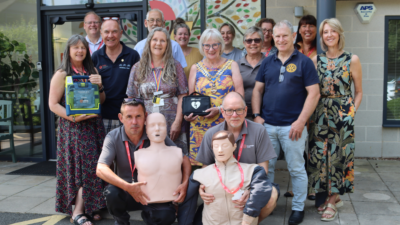 A diverse group of people stand together outdoors, some holding a defibrillator and CPR training mannequins, smiling in front of a building.