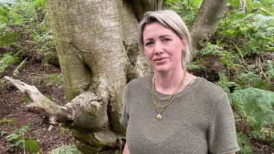 A person with short, light hair stands in front of a large tree, surrounded by lush green foliage and ferns.