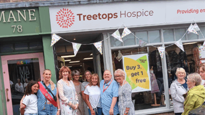 A group of people stands outside Treetops Hospice, with a "Buy 3, get 1 free" offer visible in the store window. Colorful bunting is hanging above.