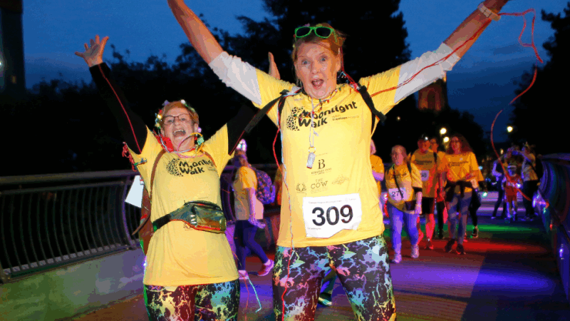 A vibrant night walk event features participants in yellow shirts and colorful leggings celebrating together under bright lights.