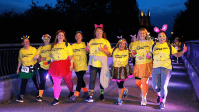 A group of cheerful participants in bright yellow shirts and colorful accessories walk together at night during a charity event.