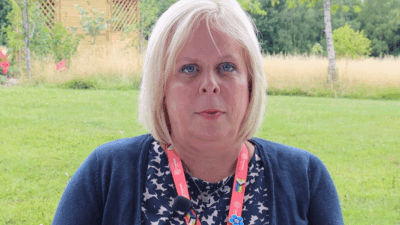 A woman with blonde hair, wearing a navy cardigan and a colorful lanyard, stands outdoors with greenery and a wooden structure in the background.