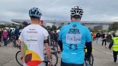 Two cyclists in kits stand among a crowd at a cycling event, one wearing a "Team Treetops" jersey and the other a blue shirt with "For Ivan."