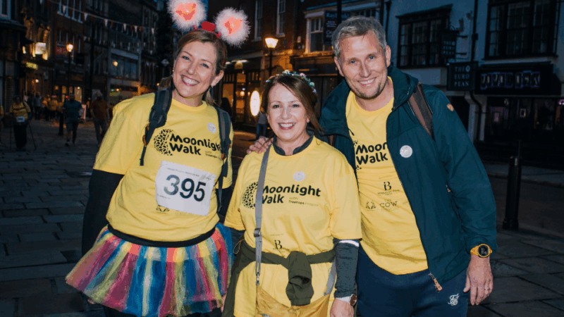 Three participants in yellow "Moonlight Walk" t-shirts stand together in a lively urban setting, wearing playful accessories and smiling.