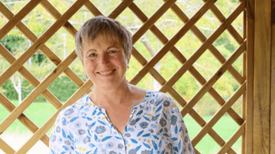 A woman with short hair wears a floral-patterned blouse, standing beside a wooden lattice structure with a green backdrop.