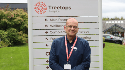 A staff member stands in front of the Treetops Hospice sign, showcasing various services while surrounded by greenery and a hospice building.