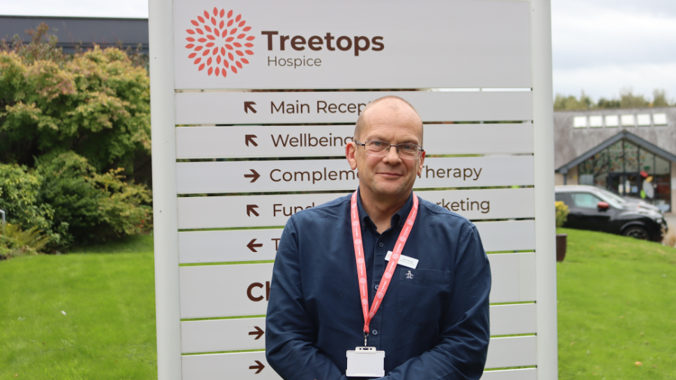 A staff member stands in front of the Treetops Hospice sign, showcasing various services while surrounded by greenery and a hospice building.