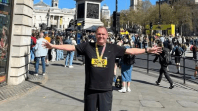 A man in a black shirt with a medal stands with arms outstretched in front of a crowded street festival on a sunny day.
