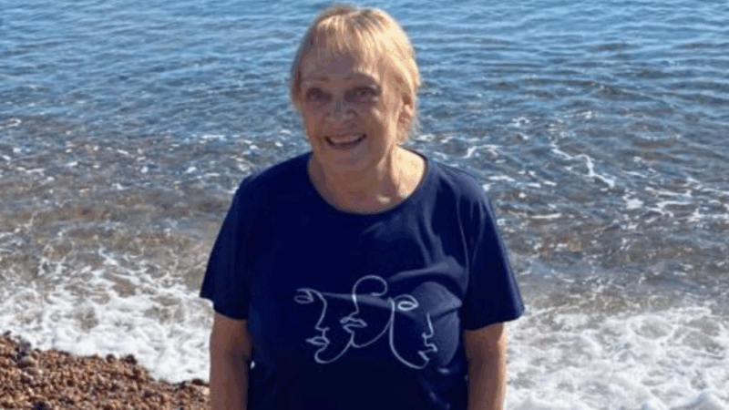 A woman stands barefoot on a pebble beach, dressed in a navy shirt with a graphic design, with the calm ocean in the background.