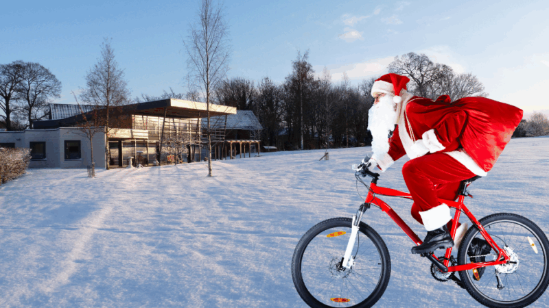 Santa Claus in a red suit rides a bicycle through a snowy landscape, carrying a sack, with a modern house in the background.