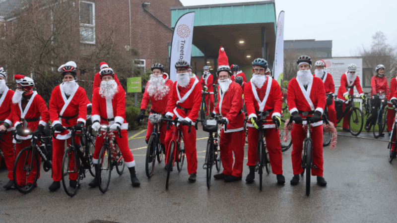 A group of cyclists dressed in red Santa costumes pose with their bikes outside a building, ready for a festive ride.