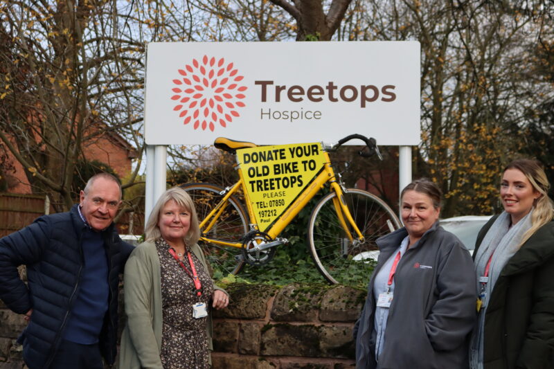 Four smiling people stood in front of a sign that reads 'Treetops Hospice'. The sign has a bike in front of it with a sign on it that reads 'donate your old bikes'.