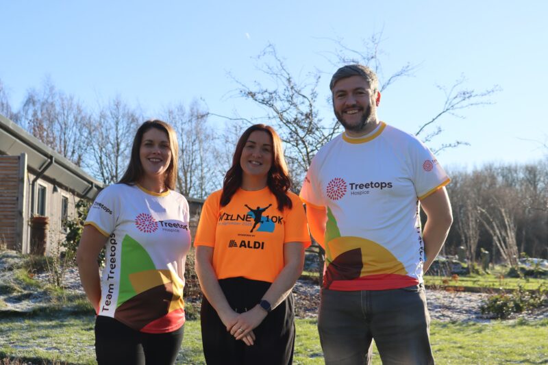 Two women and a man stood together outside in the sun smiling with T-shirts that read 'Treetops' and 'Wilne 10 k'.
