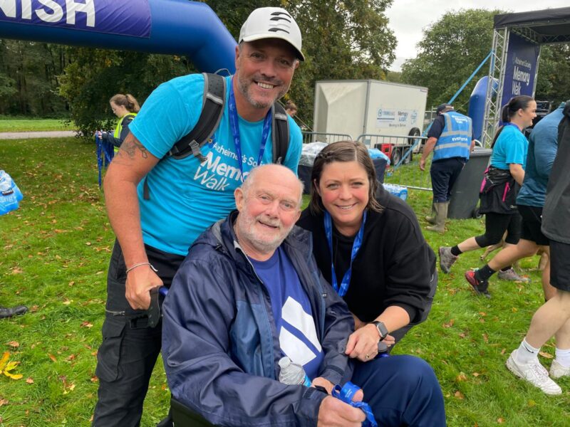 Three smiling people huddled together in a group, outside on some grass.