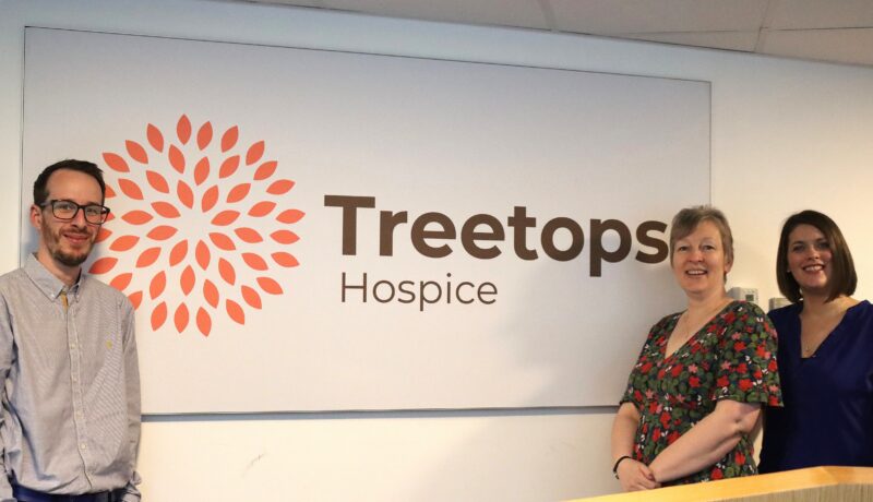 Three people smiling, next to a sign that reads 'Treetops Hospice'.