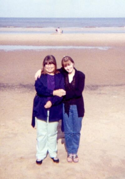 Two women smiling, arm in arm on the beach.