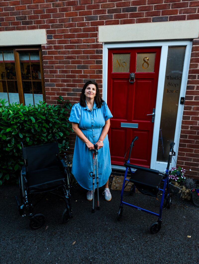 A woman stood outside of a house with her walking aids and wheelchair next to her.