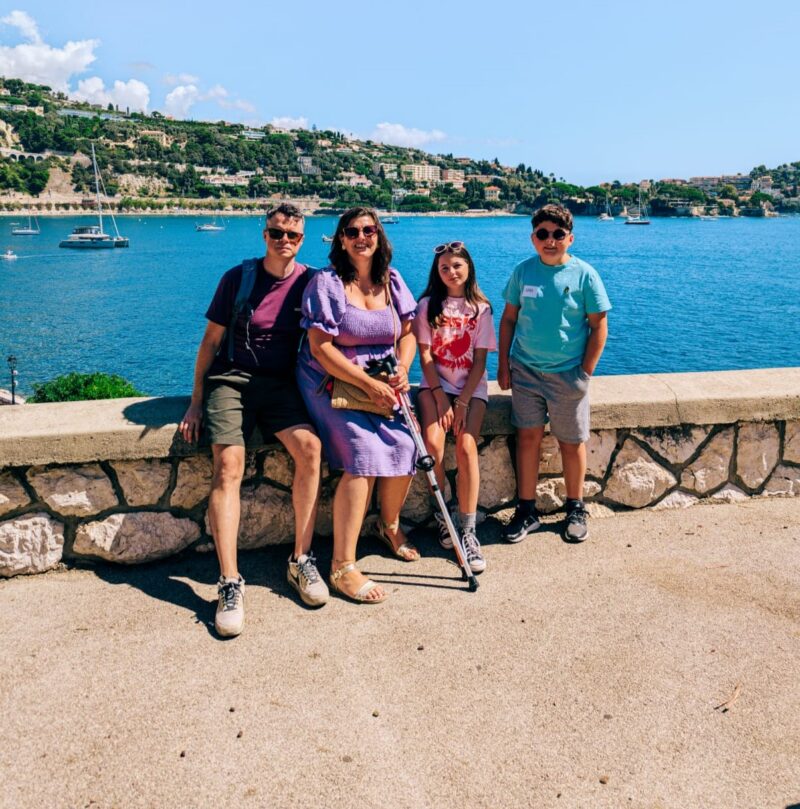 A family with two adults and two children, sat on a wall on holiday with a beach behind them.