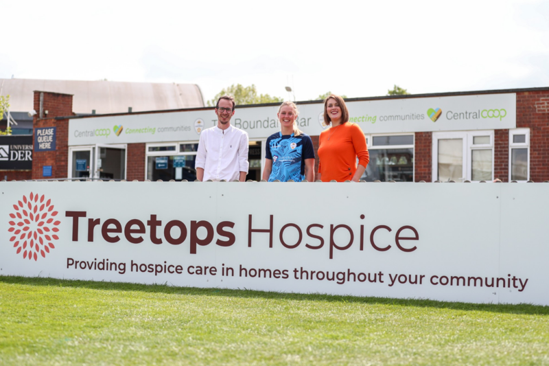 A man and two women stood behind a big board on a cricket pitch that reads 'Treetops Hospice'.