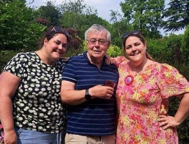 Two women smiling with a man stood in in between of them, who is also smiling.
