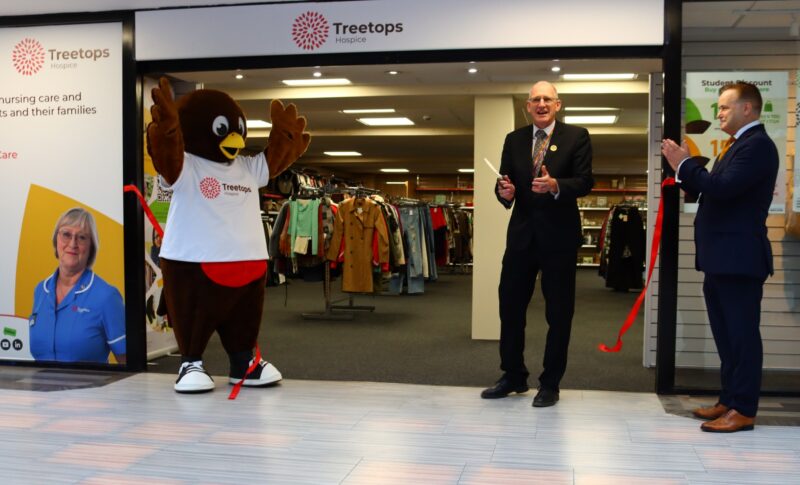 Two men smiling and clapping as a ribbon is cut and Remy, the Treetops mascot is cheering.