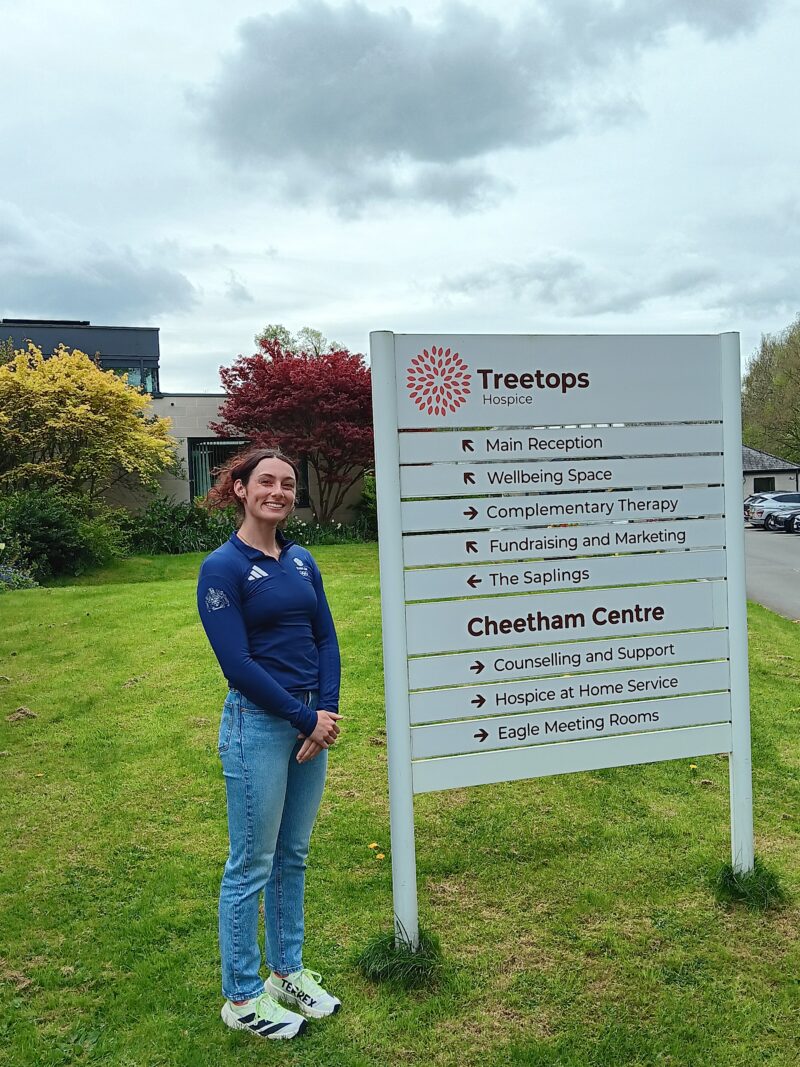 A woman smiling next to the Treetops Hospice sign.